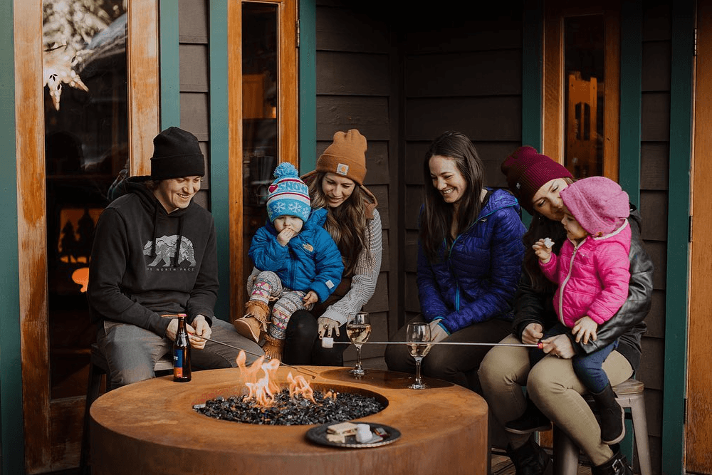 A family gathers around a fire pit, roasting marshmallows and enjoying drinks on a cozy winter evening.