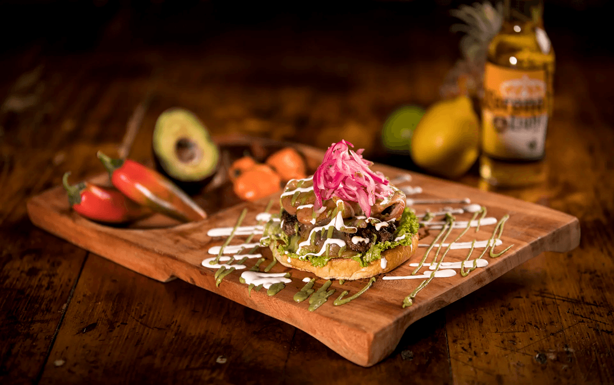 A delicious burger topped with pickled onions, served on a wooden platter alongside fresh peppers and an avocado.