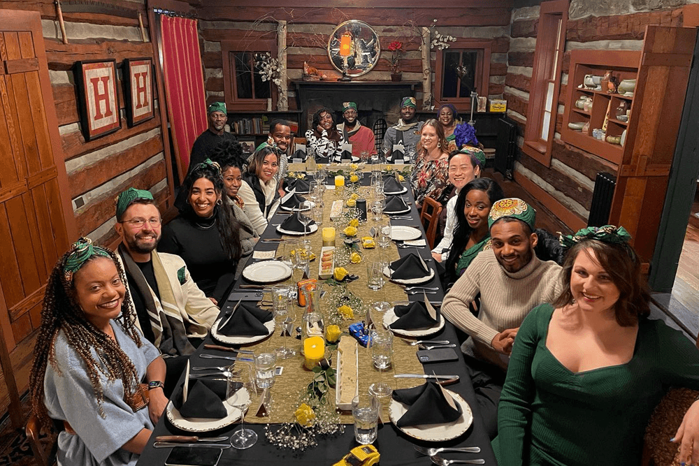 A group of diverse individuals seated around a decorated dining table in a cozy cabin setting, all wearing colorful head wraps.