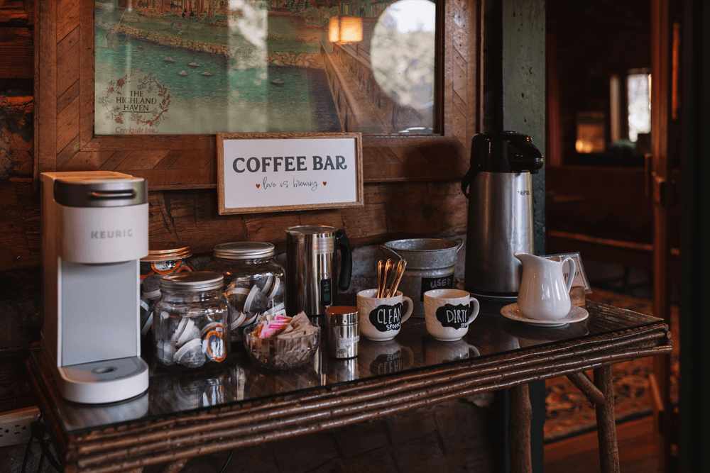 A cozy coffee bar setup with a Keurig machine, jars of coffee pods, and labeled mugs on a rustic table.