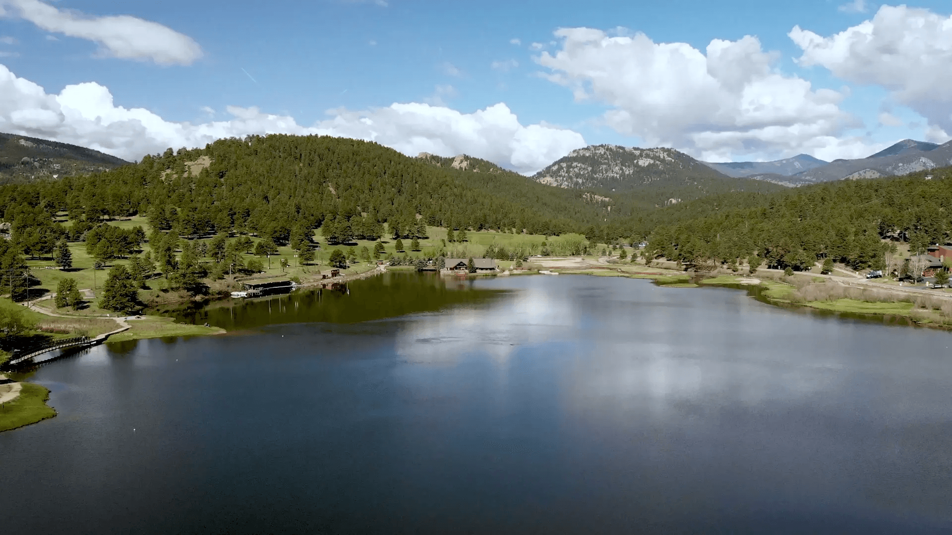 A serene lake reflects the blue sky and white clouds, surrounded by lush green hills and distant mountains under a bright, clear day.