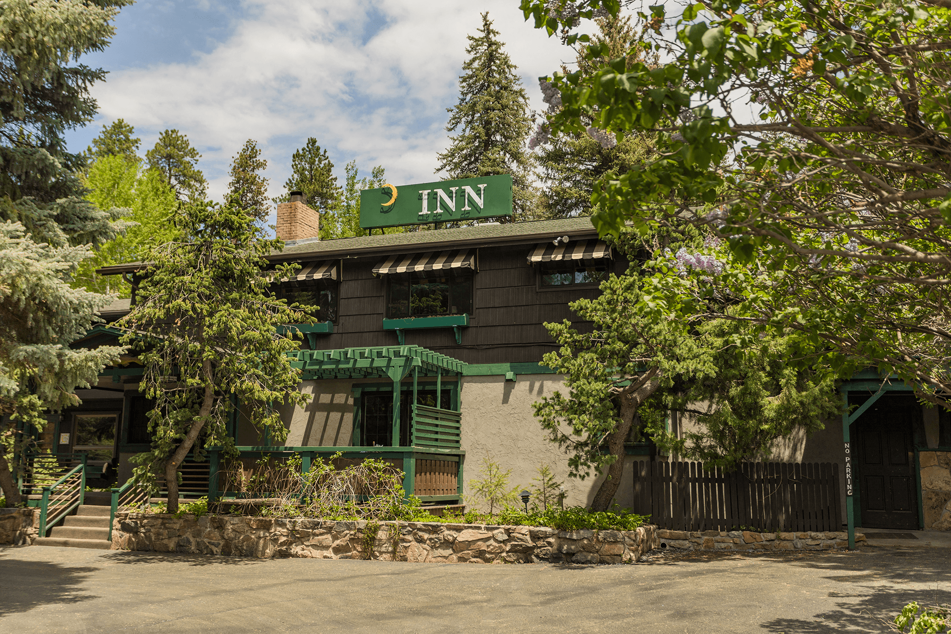 Two-story rustic inn surrounded by lush green trees, featuring a stone base, a wooden porch, and a forest backdrop, with a relaxed, inviting tone.