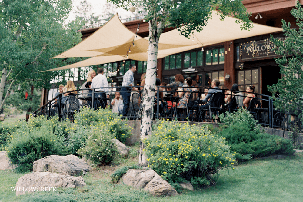 People gather on a patio under shade sails surrounded by greenery.