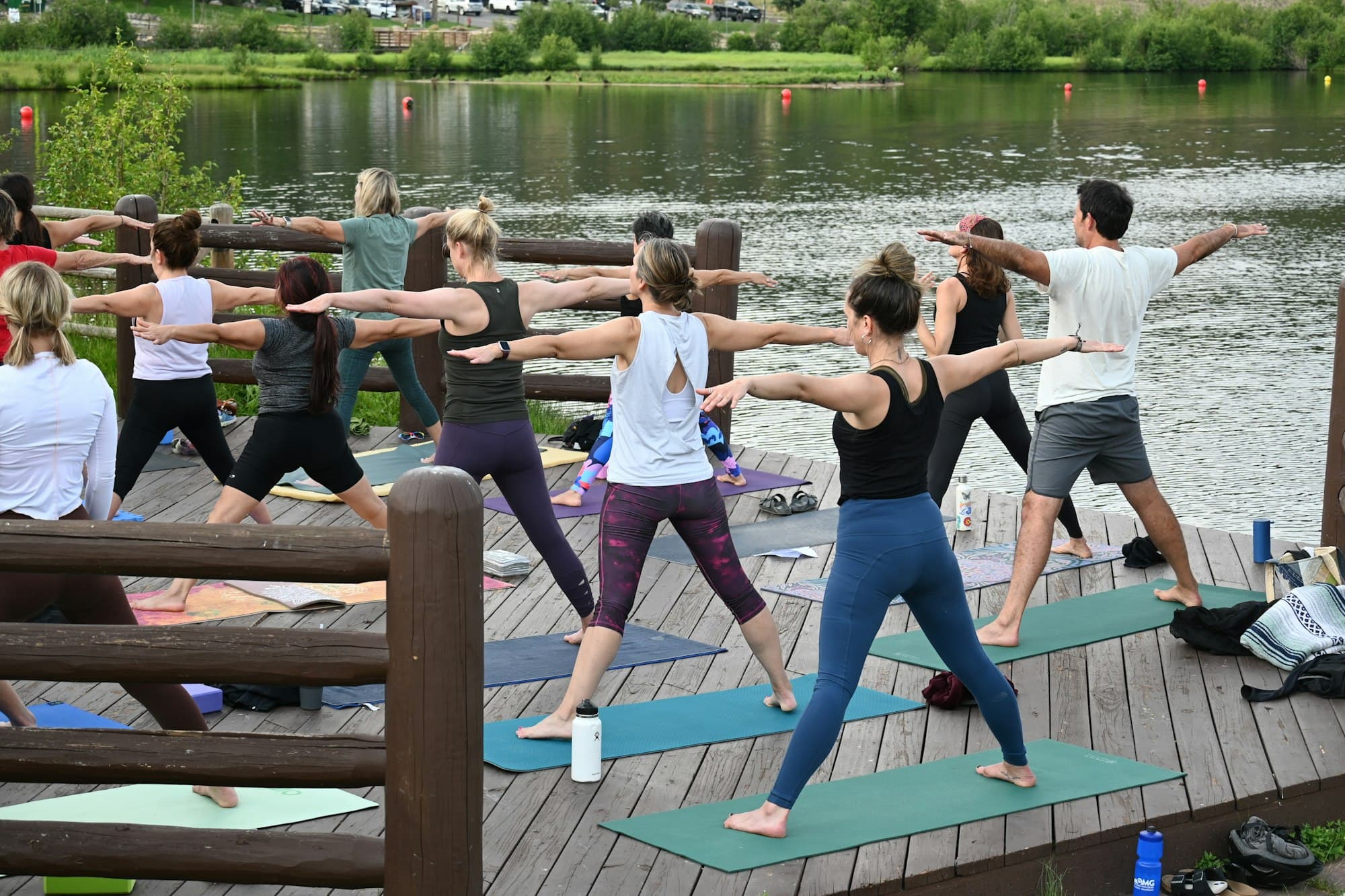 A group of people practice yoga on a wooden deck by a serene lake, all in the Warrior II pose. The setting is calm with lush greenery in the background.