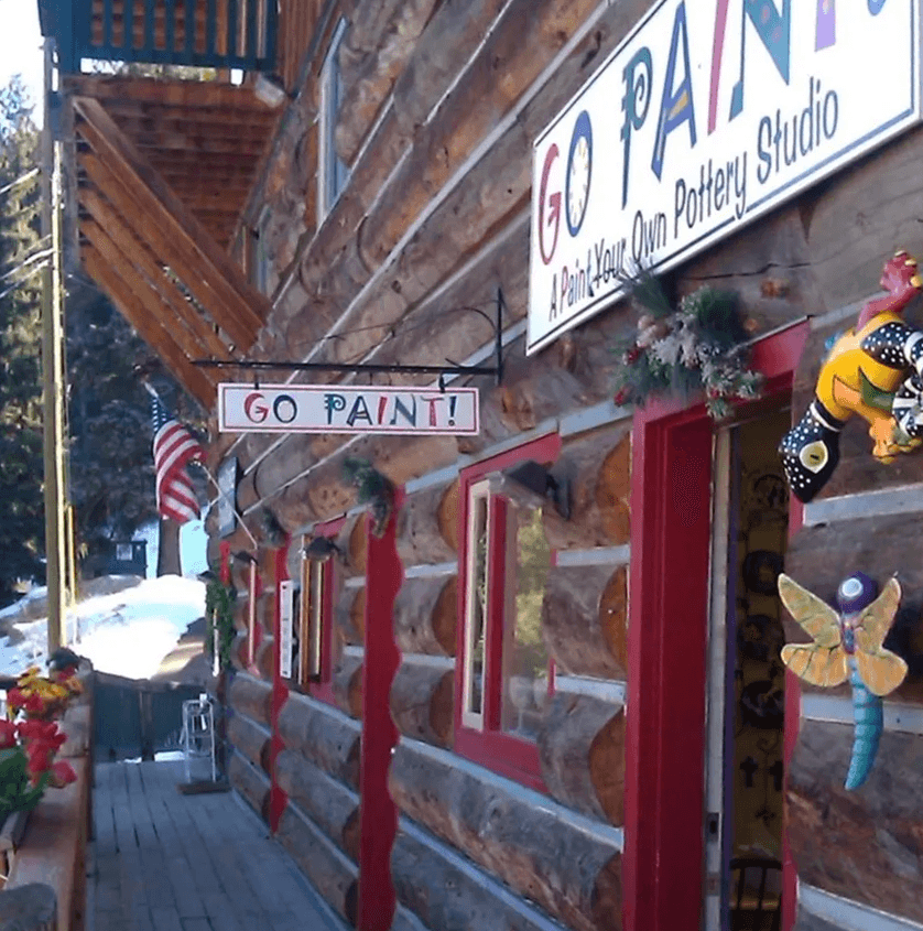 A log cabin pottery studio with a colorful sign that reads "GO PAINT!" and decorative ornaments.