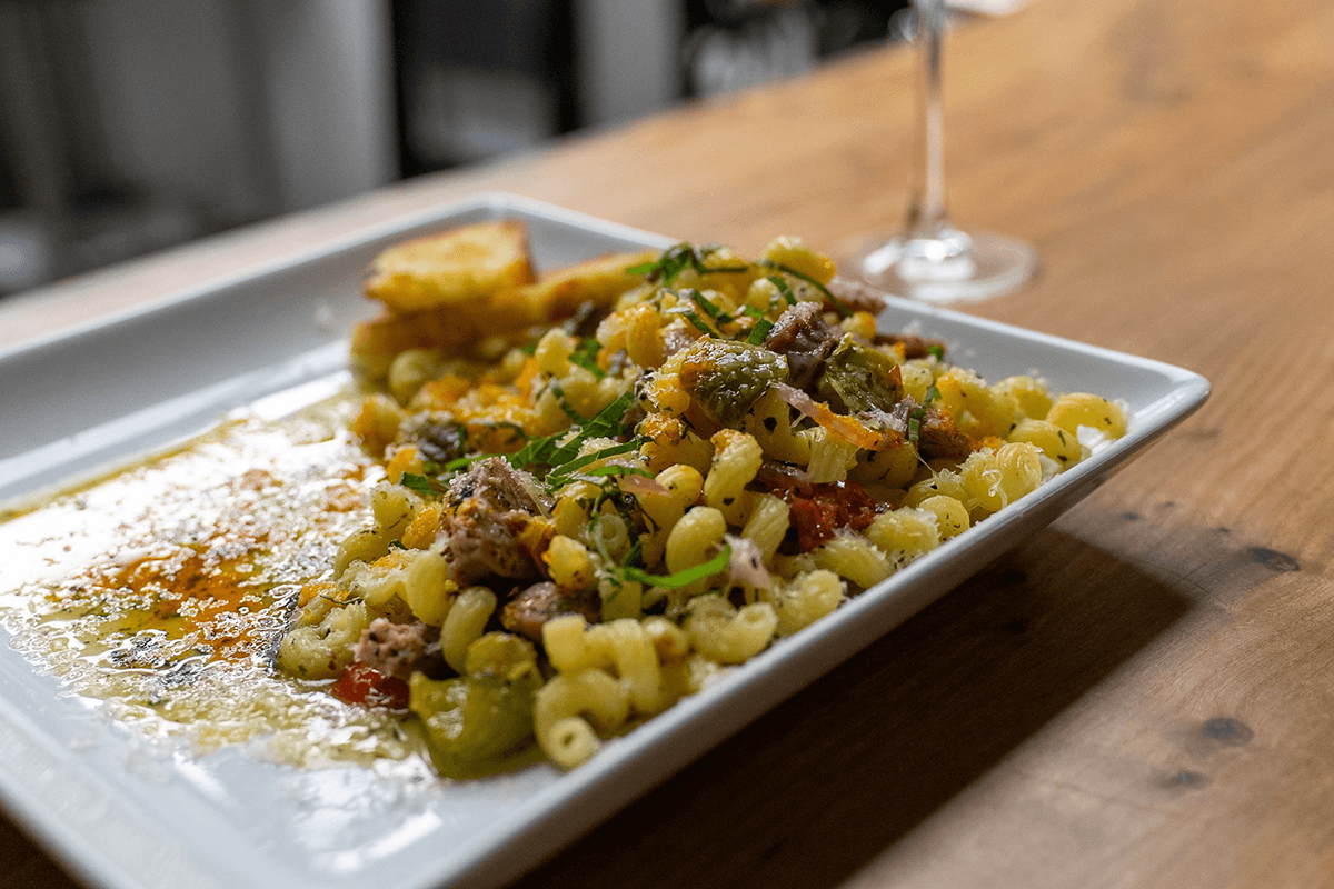 A close-up of pasta with vegetables and meat served on a white plate, alongside a piece of bread and a glass of wine in the background.