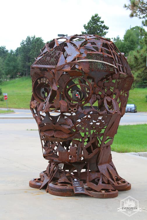 A large, abstract metal sculpture resembling a human head stands outdoors, surrounded by greenery.