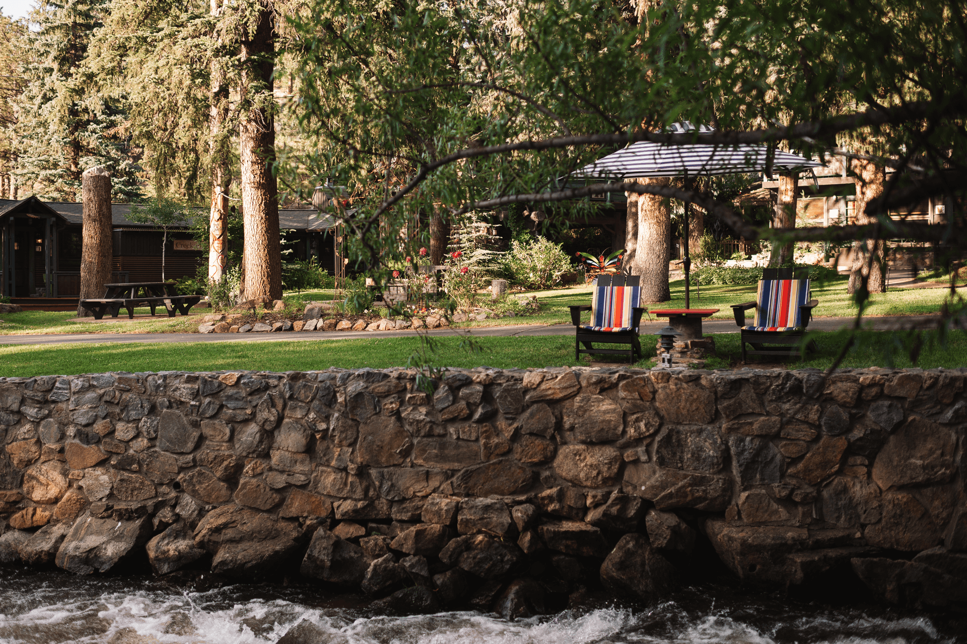 A serene outdoor scene featuring two colorful chairs by a stone wall, overlooking a river and surrounded by greenery.