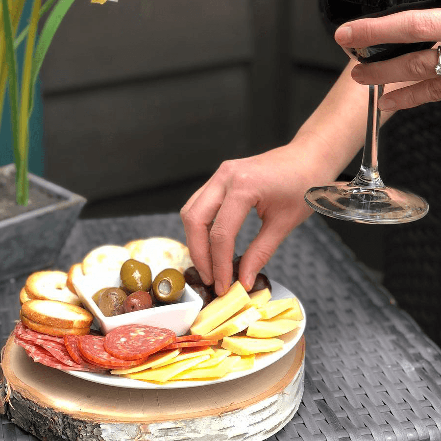 A hand reaches for a cheese and charcuterie platter accompanied by a glass of red wine.