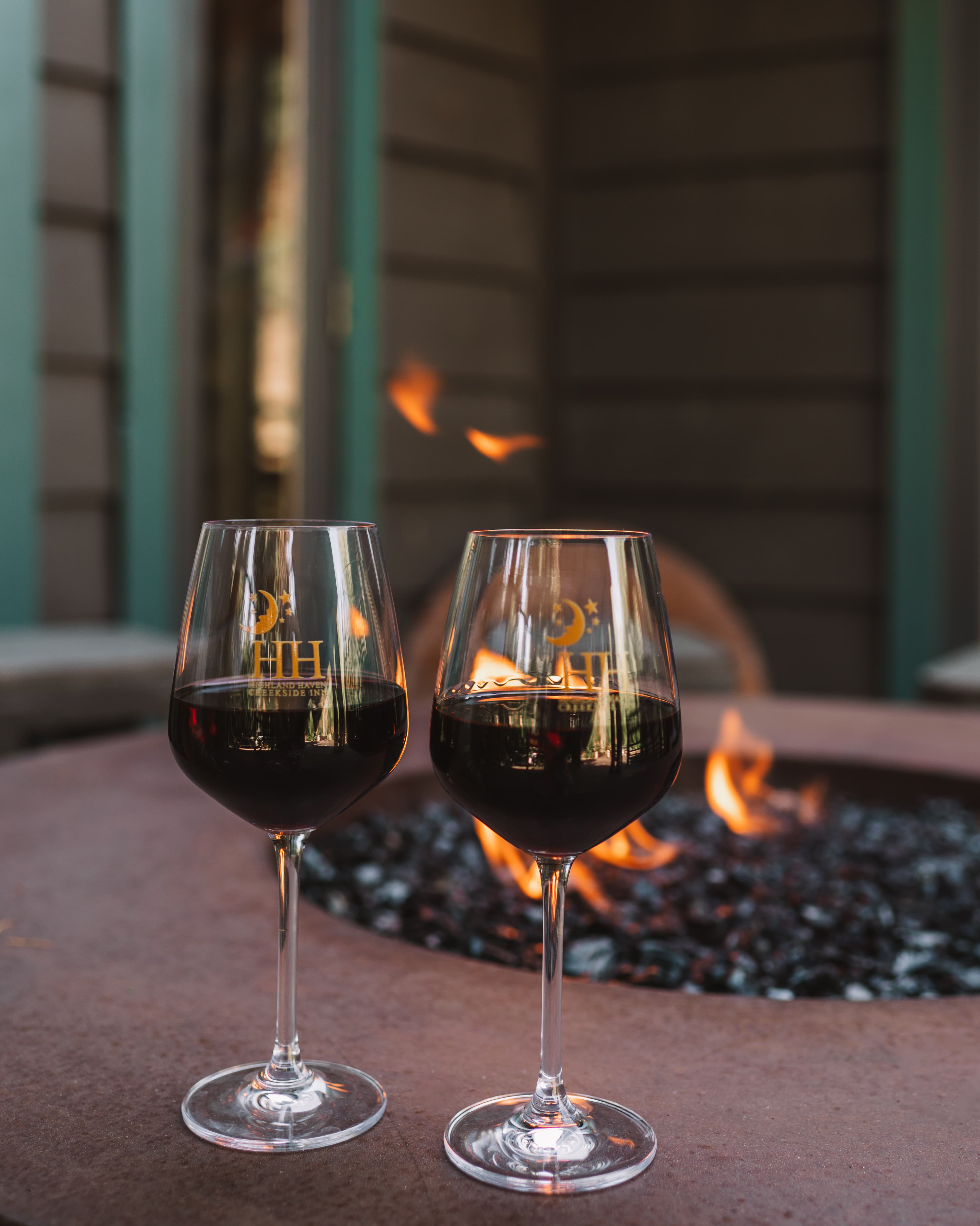 Two glasses of red wine sit on a stone table next to a fire pit.