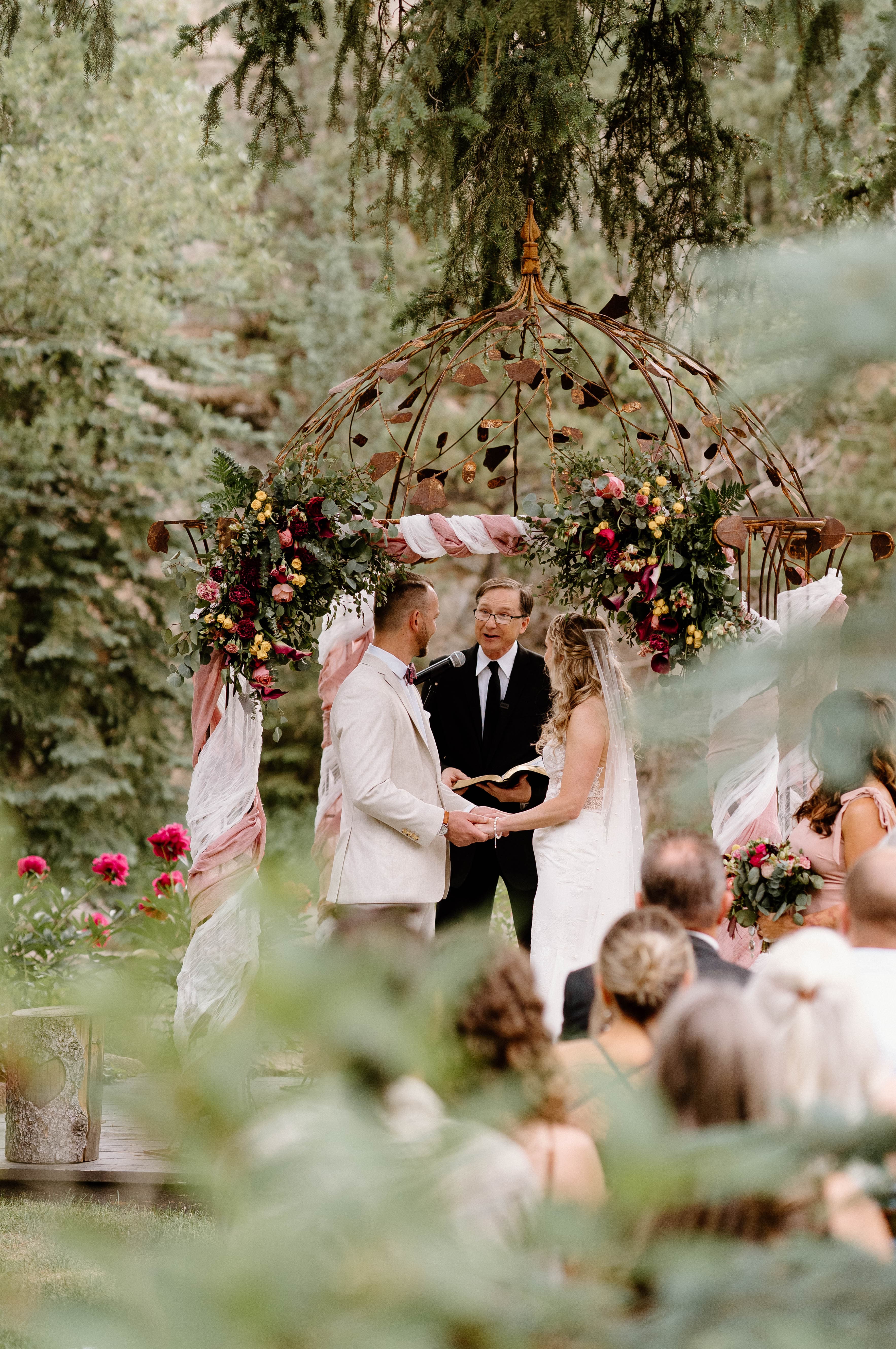 A couple exchanges vows under a floral arch during an outdoor wedding ceremony.
