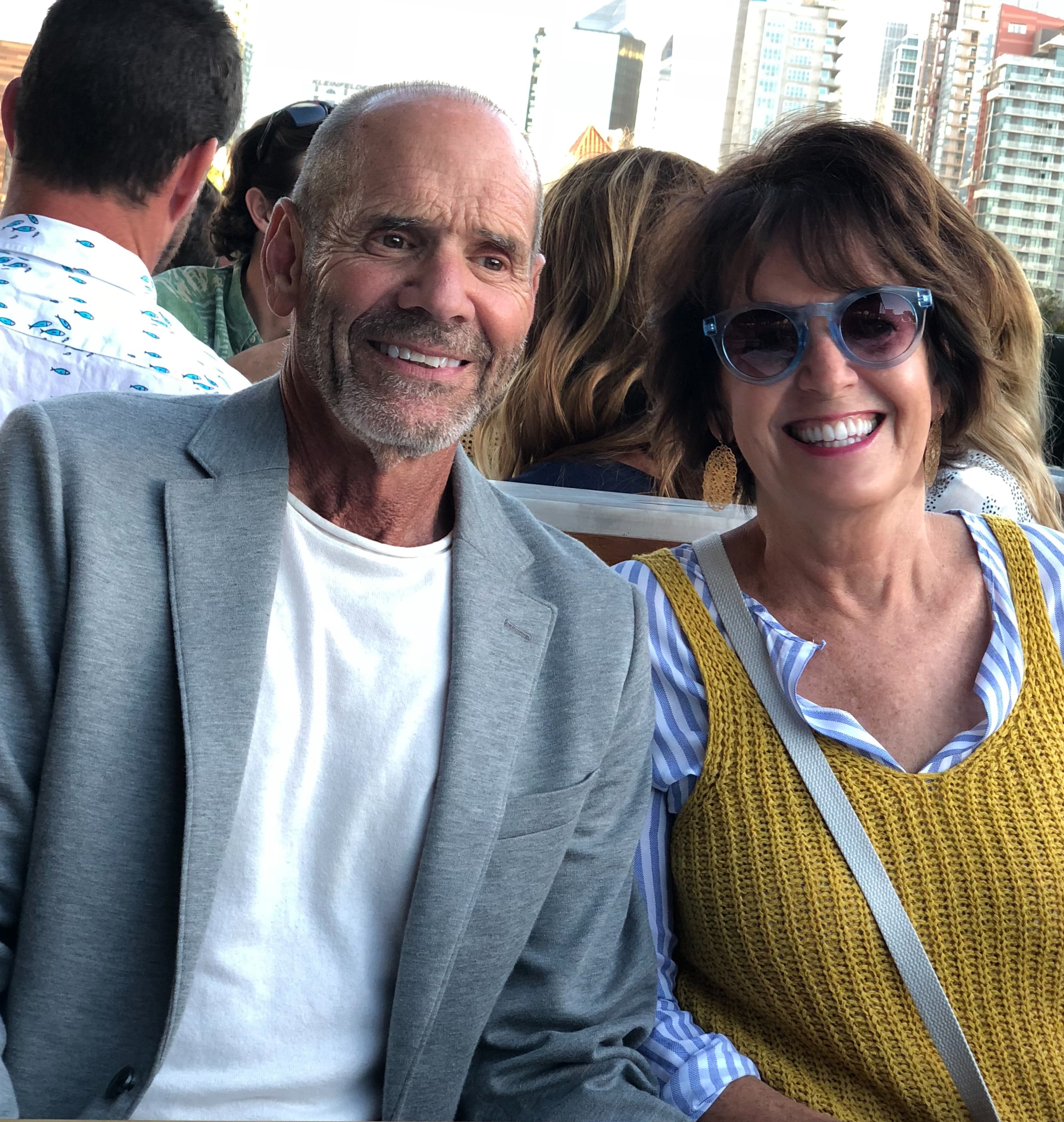 A smiling man in a gray blazer sits next to a woman in a yellow top, both enjoying a lively outdoor gathering.