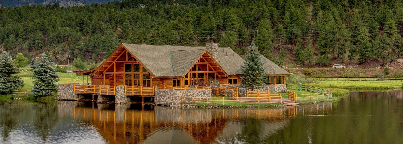 A rustic wooden house with a stone foundation sits by a serene pond surrounded by lush greenery.