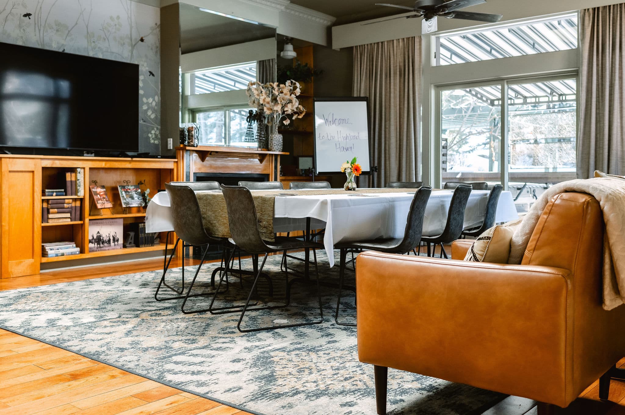 A cozy dining space featuring a long table, wooden shelves, and a large TV, with sunlight streaming through the windows.