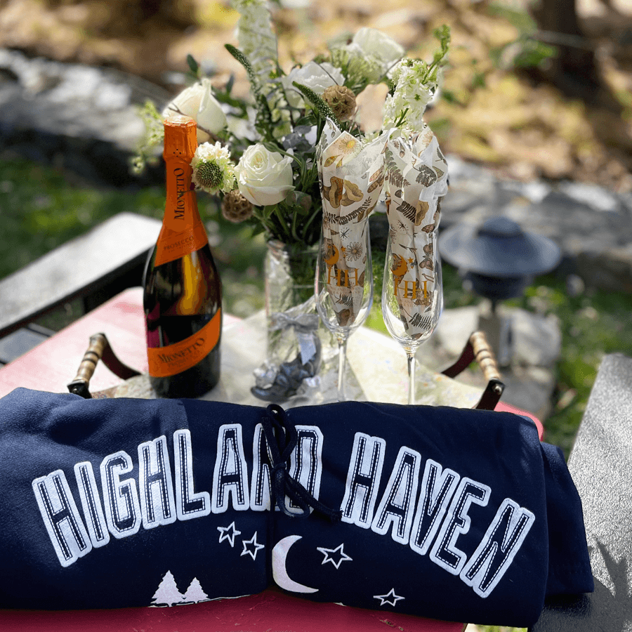 A navy sweatshirt with "HIGHLAND HAVEN" printed on it, a bottle of Prosecco, two decorated champagne flutes, and a vase of white flowers are arranged on a table outdoors.