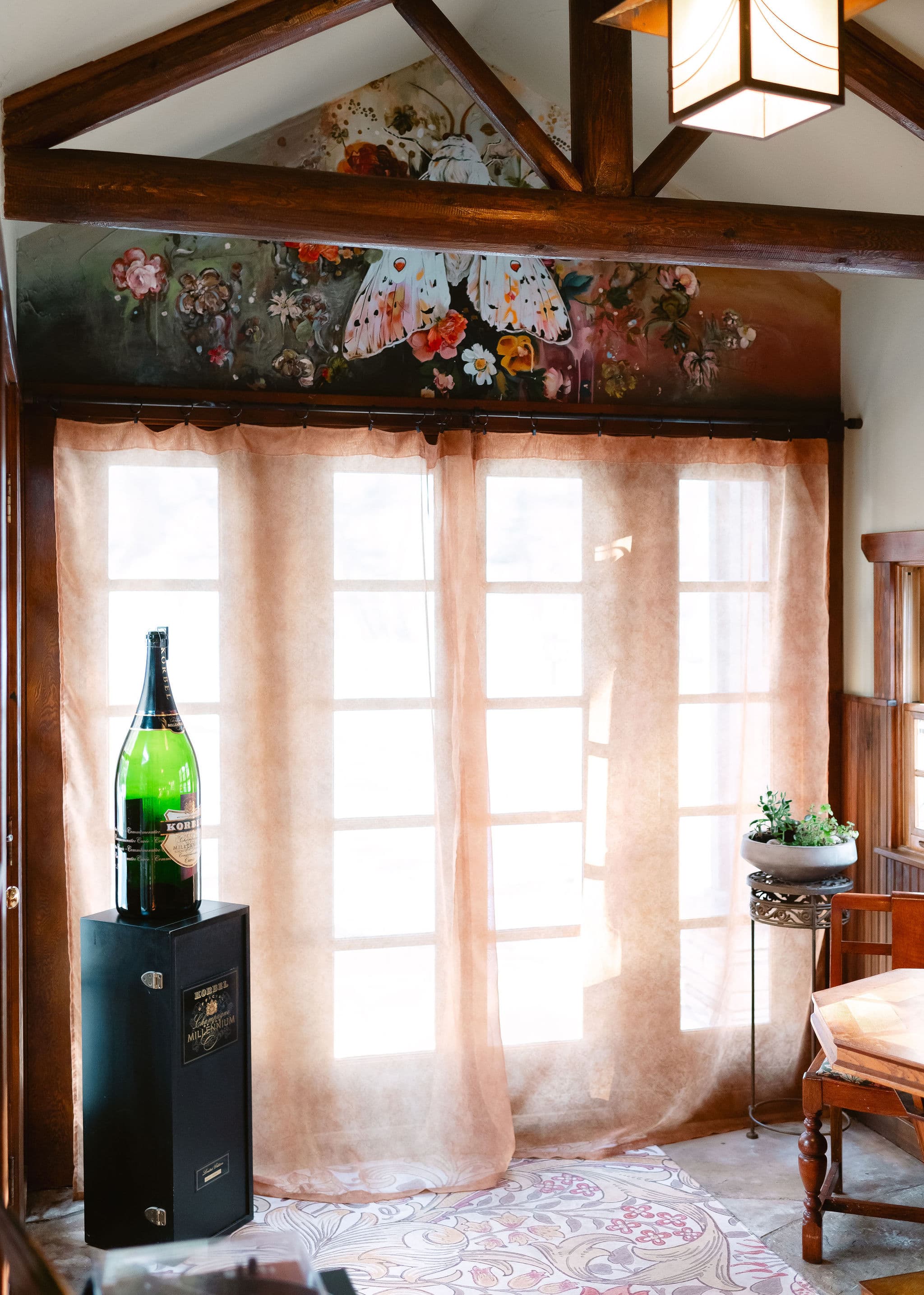 A large green bottle stands on a black display next to sheer curtains and decorative plants in a warmly lit room with floral artwork on the wall.