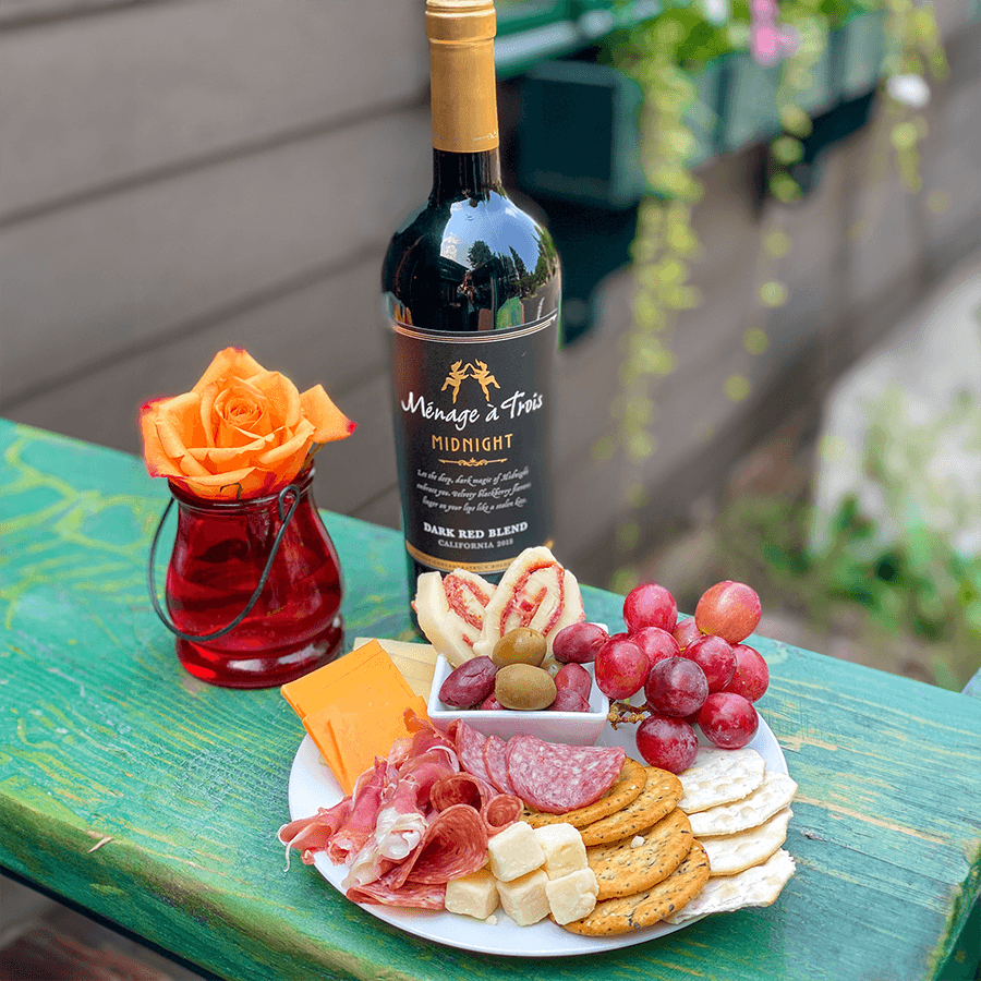 A bottle of Ménage à Trois Midnight wine sits next to a platter of assorted cheeses, cured meats, crackers, grapes, and a small vase with an orange rose.