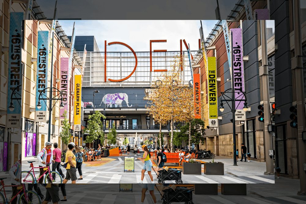 Urban street scene in Denver with colorful banners and the word "DEN" in large letters. People walk and cycle on a vibrant pedestrian street.