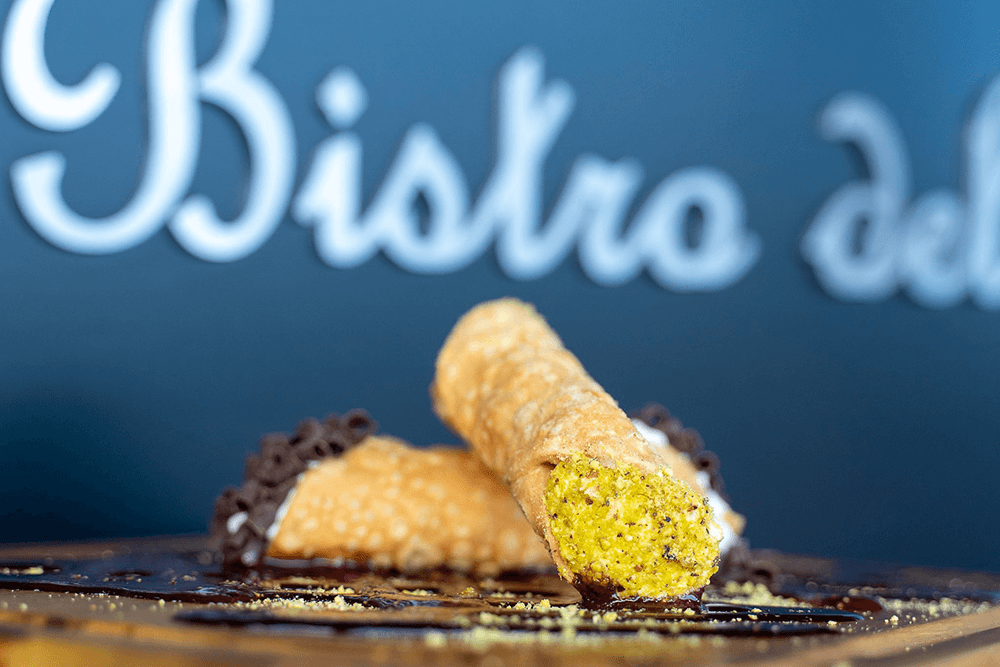 A close-up of a cannoli filled with pistachio cream, garnished with chocolate shavings and placed on a wooden surface.