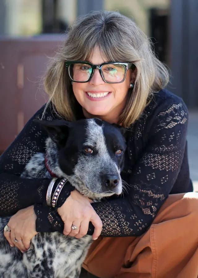 A woman with glasses smiles while hugging a black and white dog.