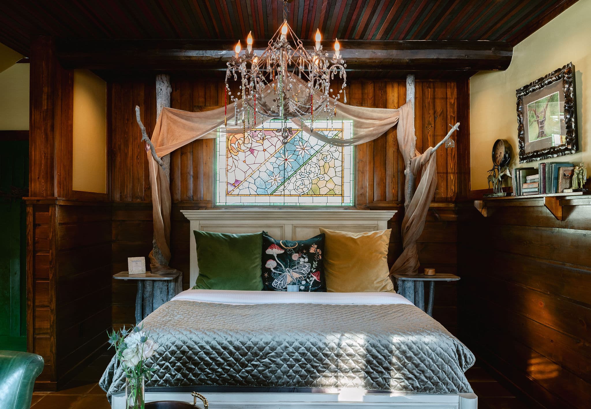 A cozy bedroom featuring a stained glass window, a chandelier, and plush pillows on a bed with a quilted cover.