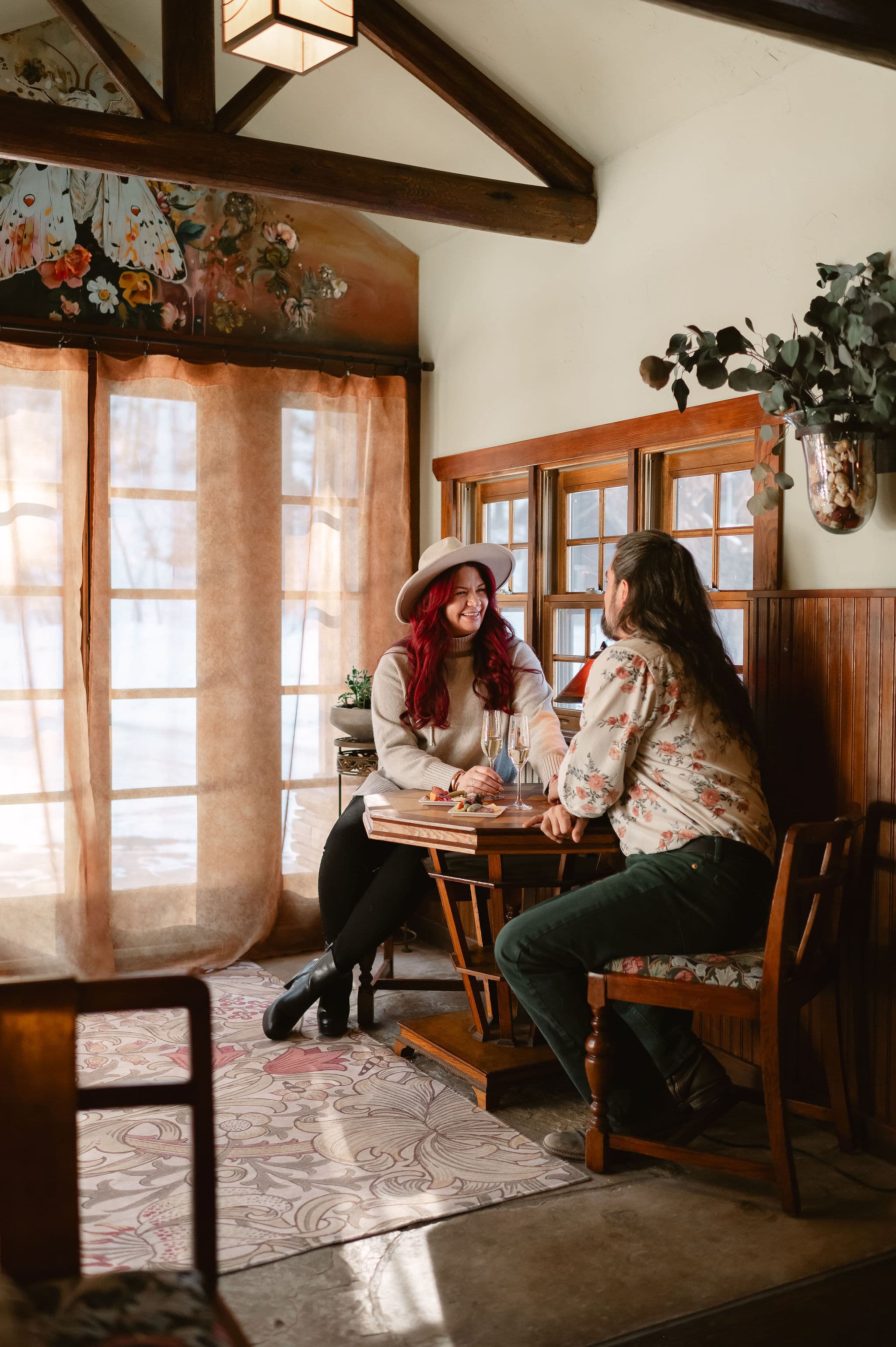 A couple enjoys a cozy conversation over drinks at a wooden table in a warmly lit room.