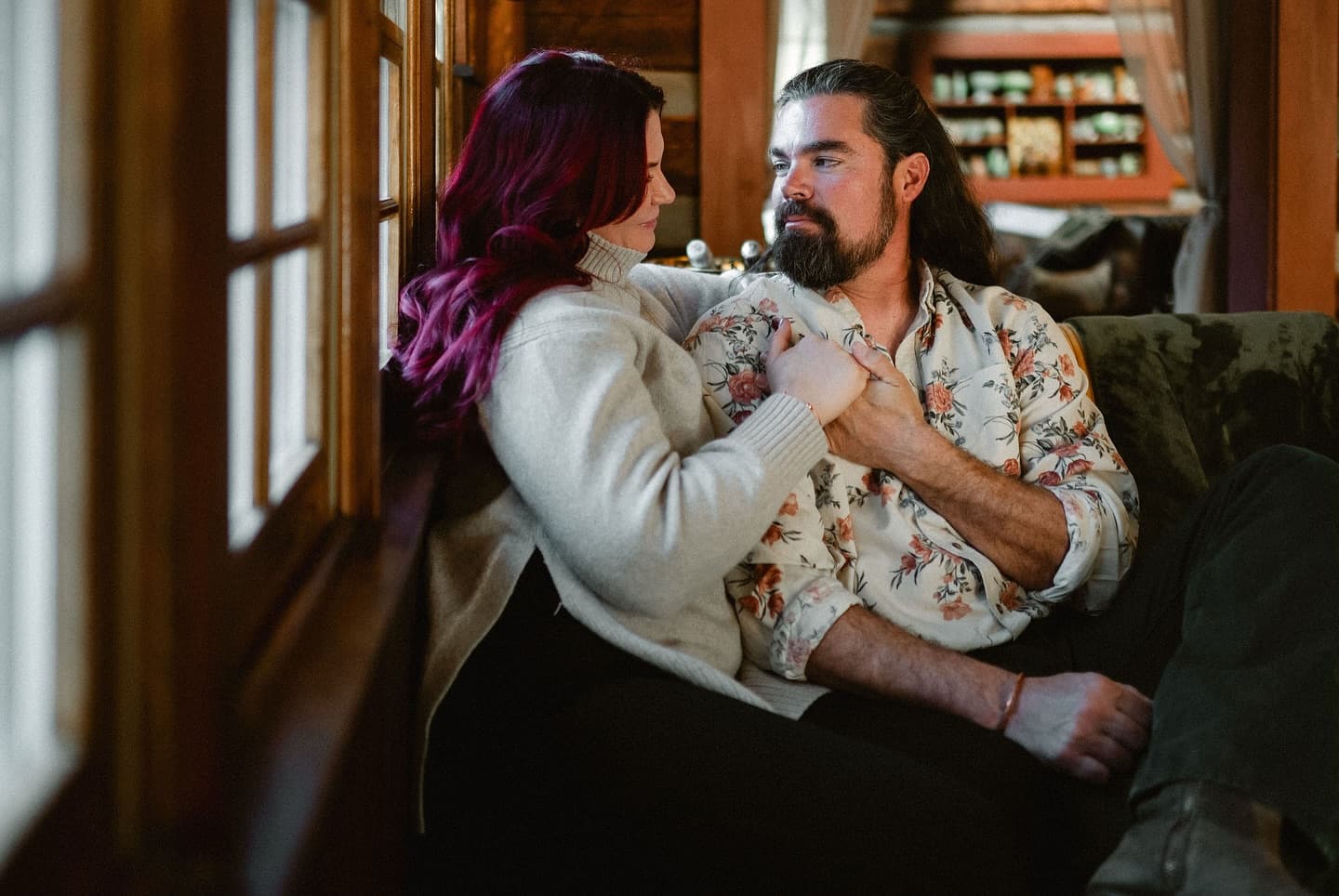 A couple sits closely on a cozy couch, gazing into each other's eyes. The room is warmly lit, with wooden paneling and shelves in the background.