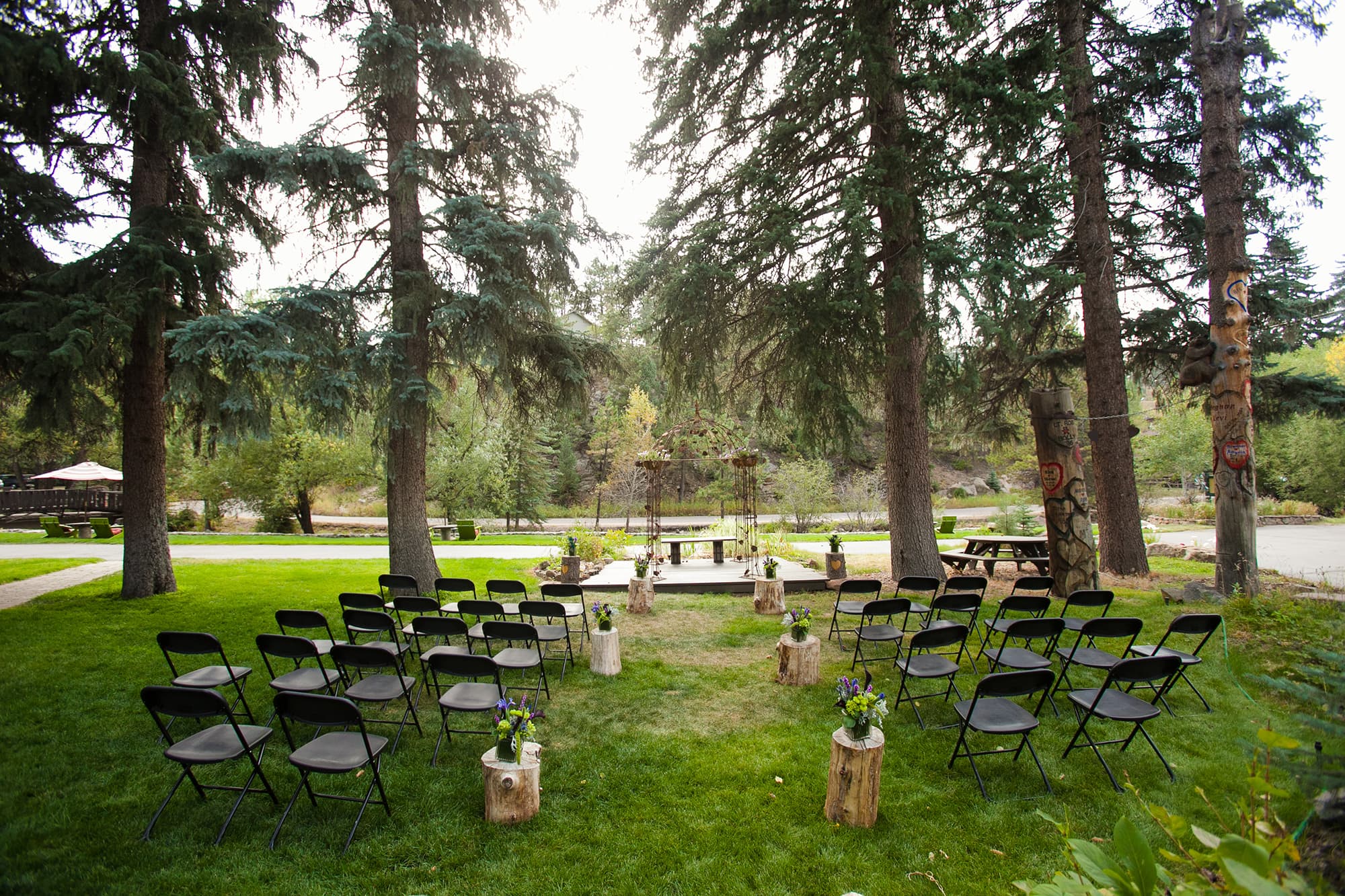 An outdoor wedding setup featuring black chairs arranged around a wooden altar, surrounded by tall trees.