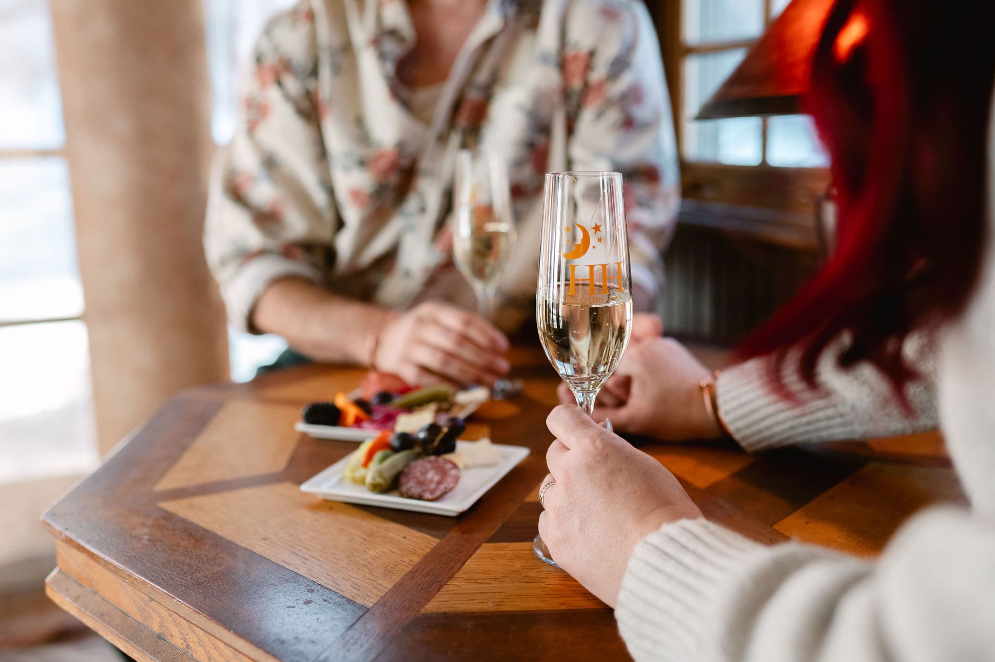 Two people are enjoying a cozy meal at a wooden table, holding champagne flutes. A small charcuterie plate adds a touch of elegance.