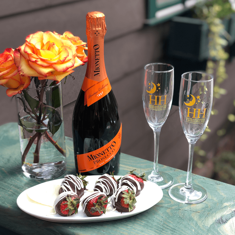 A bottle of Mionetto Prosecco, two champagne flutes, chocolate-covered strawberries, and a bouquet of orange roses on a wooden table.