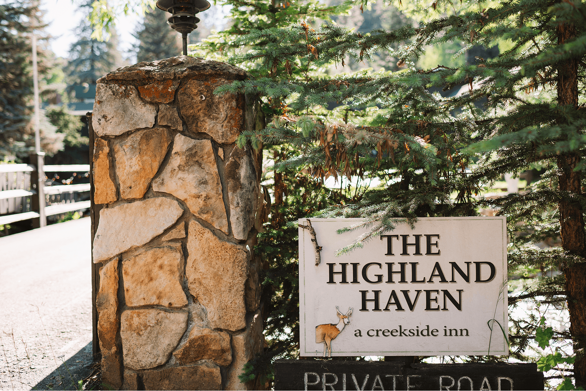 Stone pillar and evergreen tree surround a sign reading "The Highland Haven, a creekside inn" with a deer image. Sunlit, peaceful setting.