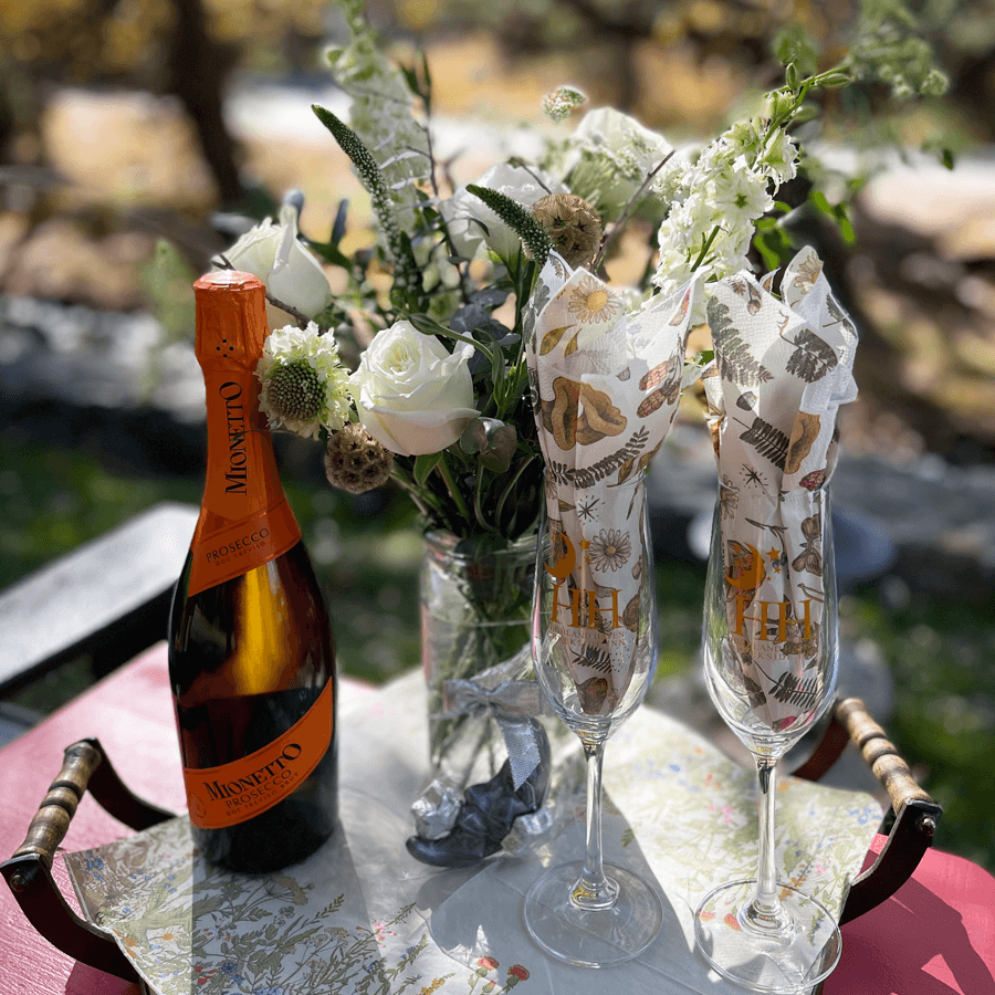 A bottle of prosecco and two wrapped champagne flutes sit on a decorative tray. A vase of white flowers adds elegance. Sunlight filters through trees, creating a serene outdoor setting.