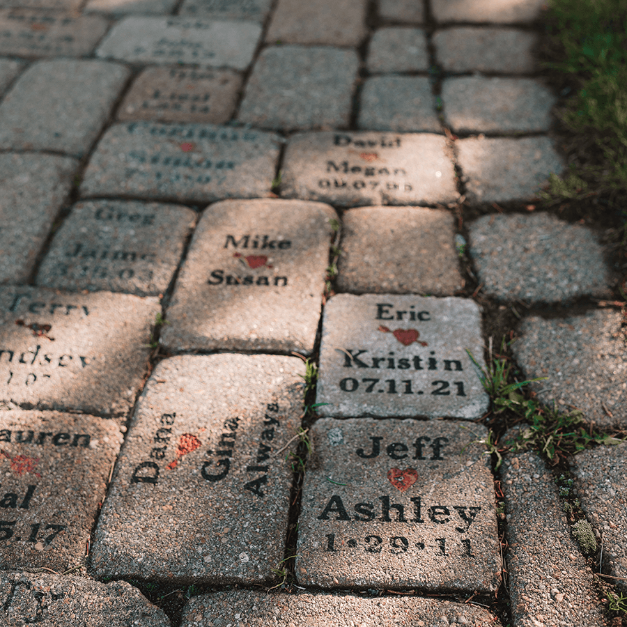 Brick path with engraved names, dates, and red heart symbols, partially shaded by sunlight with some grass peeking through gaps.