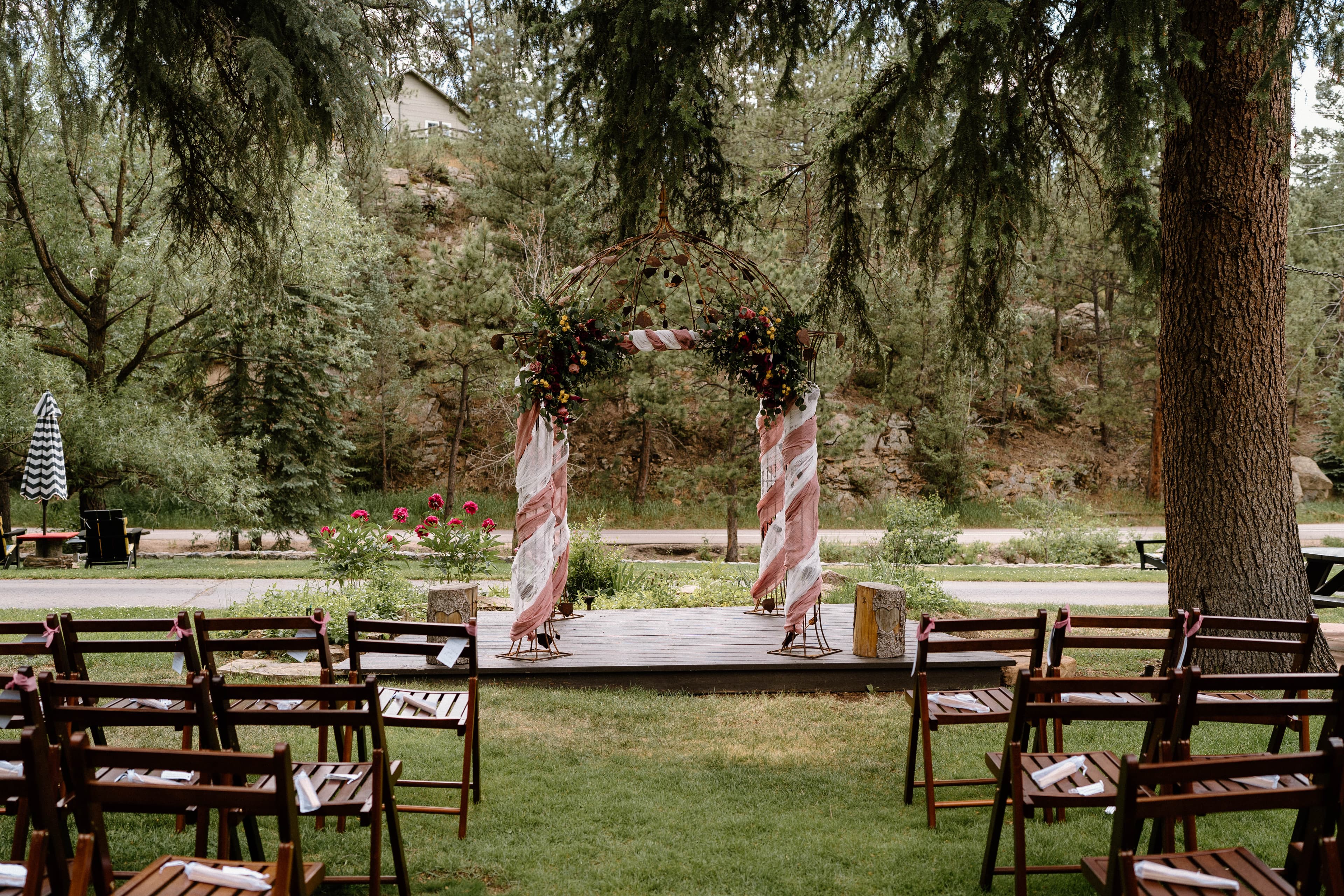 A wooden platform decorated with floral arches is framed by rows of empty wooden chairs in a lush, green outdoor setting.