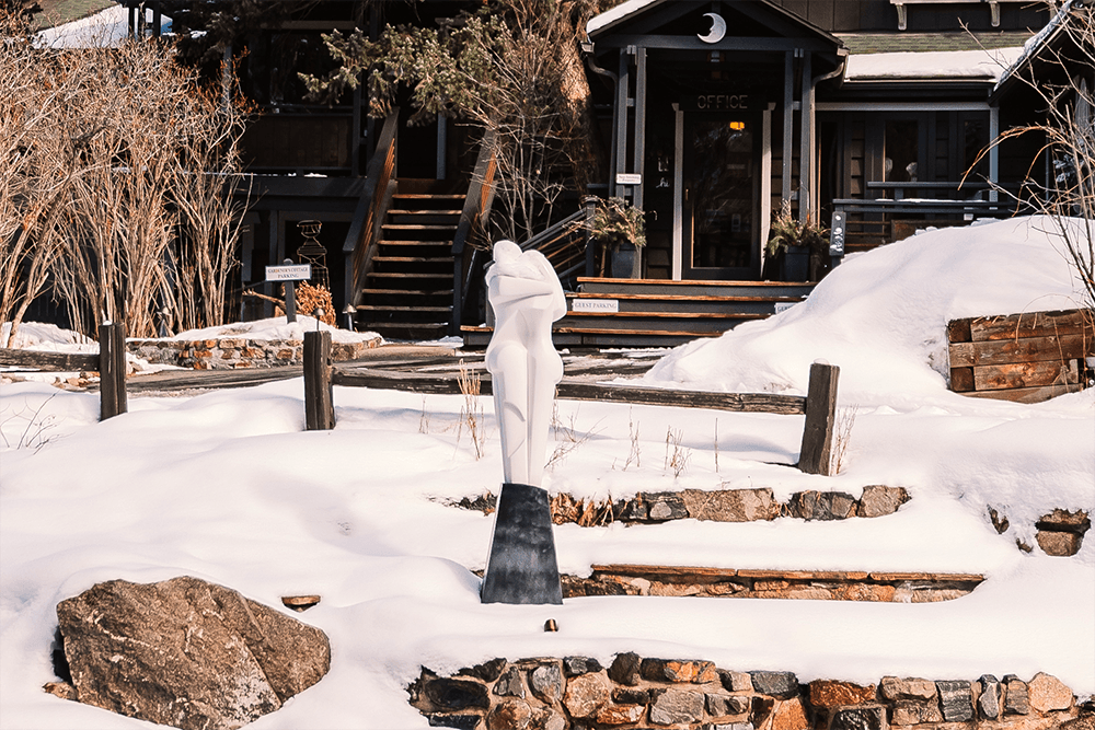 A modern white sculpture on a snowy landscape stands before a rustic wooden building. Bare trees and stone steps lead to the entrance, creating a serene winter scene.