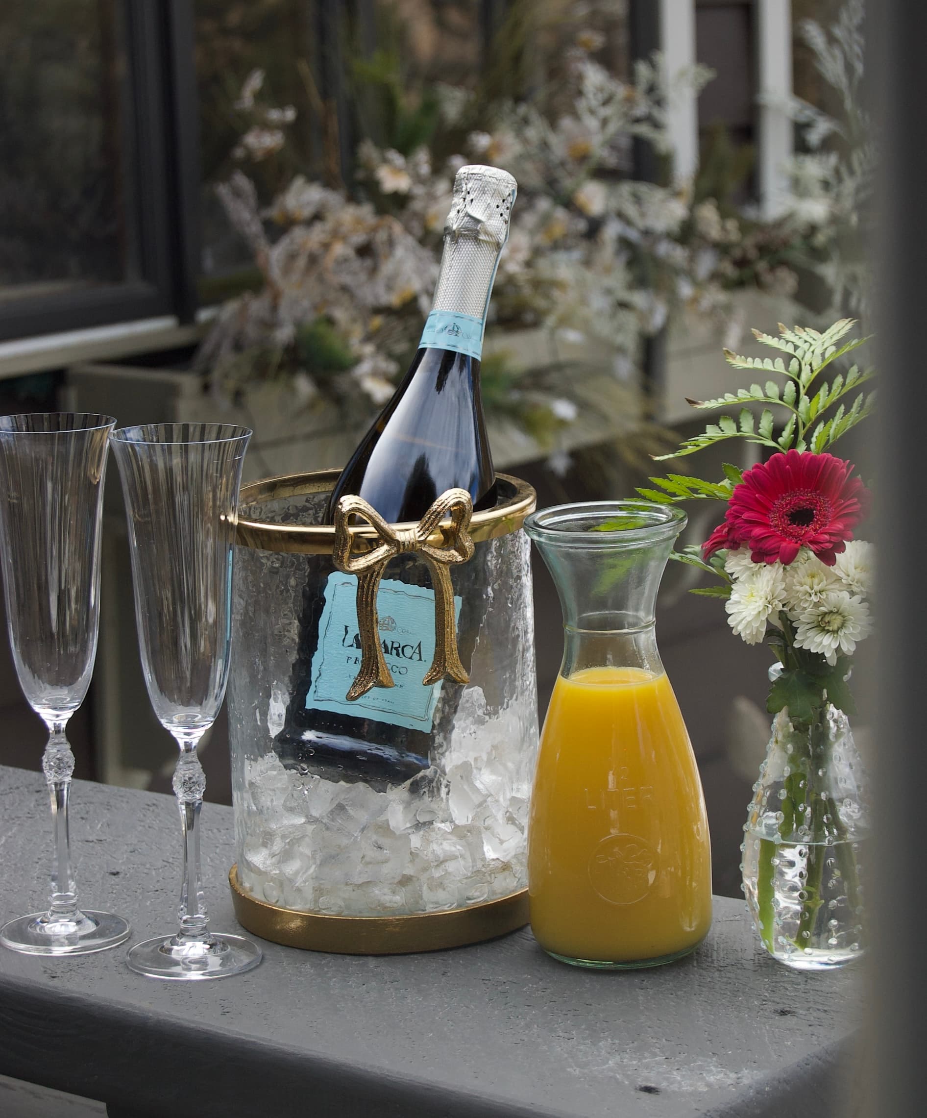 A bottle of sparkling wine in an ice bucket with two champagne flutes and a pitcher of orange juice alongside a vase of flowers.