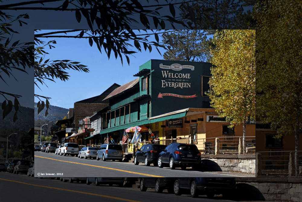 Street view of a rustic town with green wooden buildings and signage reading "Welcome to the Heart of Evergreen." Cars line the street, and flags adorn storefronts under clear blue skies.
