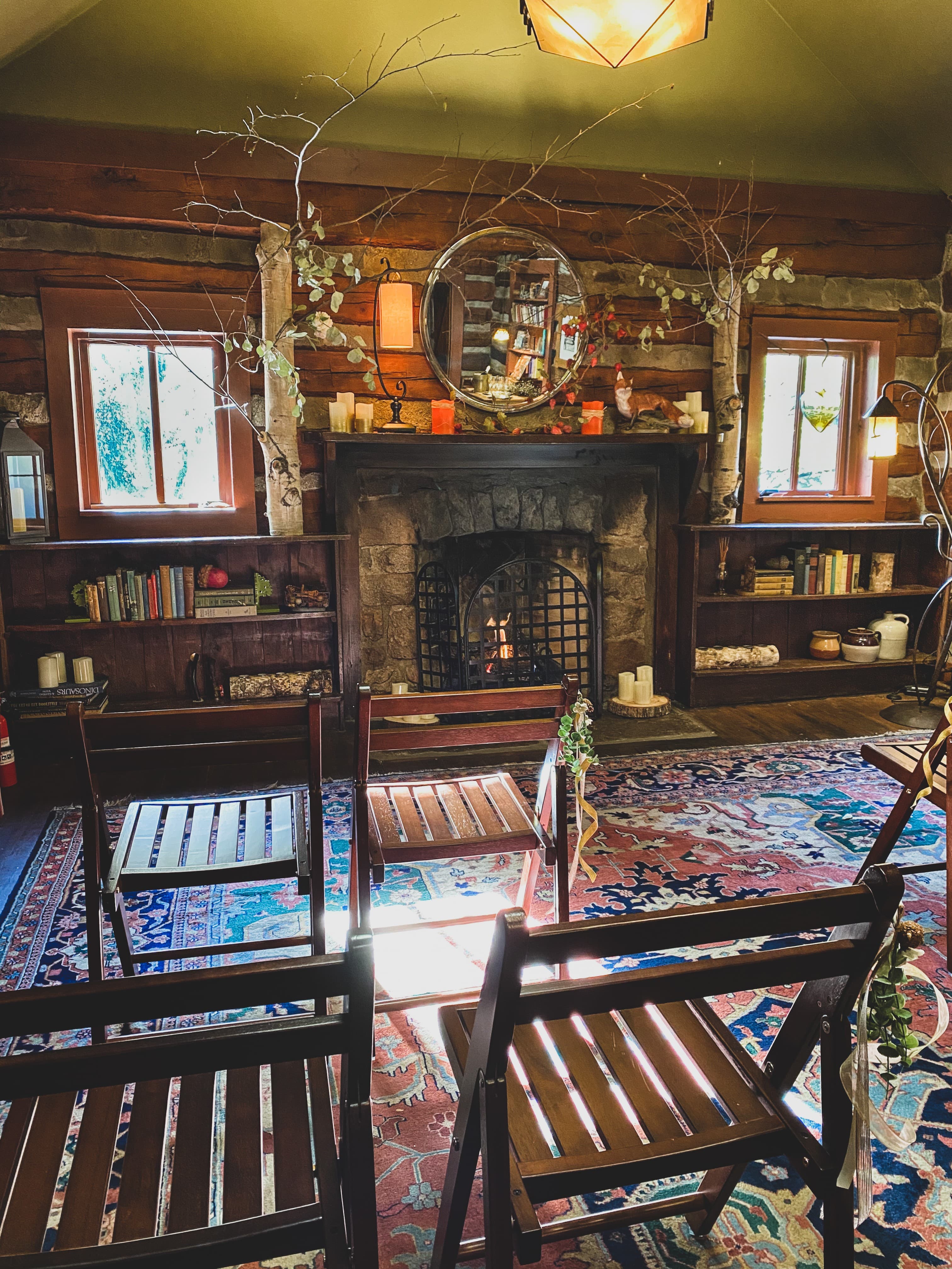 A cozy, rustic room featuring a stone fireplace, wooden shelves filled with books, and several wooden chairs arranged in front of a colorful rug.