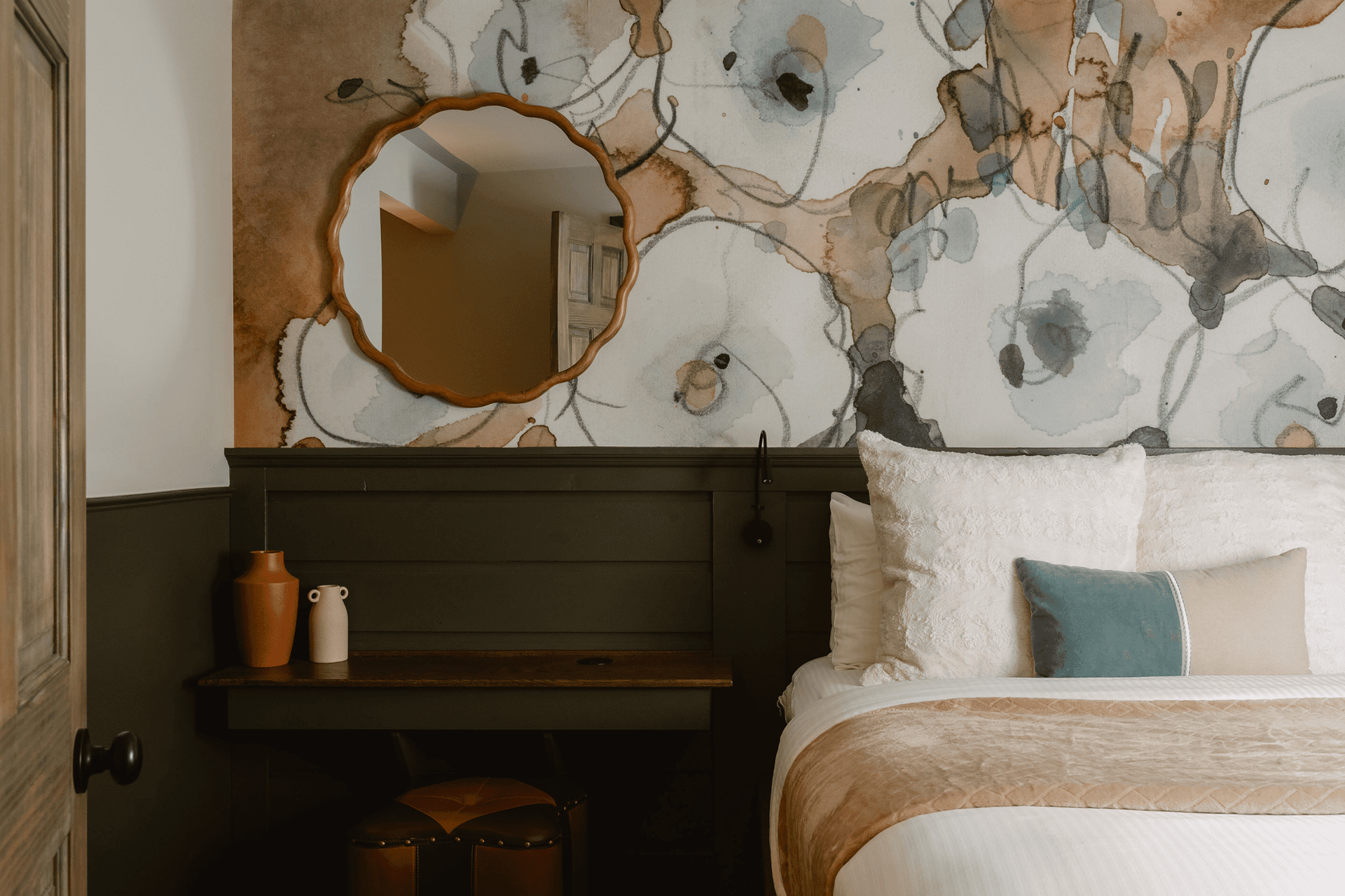 A cozy, elegantly designed bedroom featuring a floral accent wall, a round mirror, and a wooden desk with decorative vases.