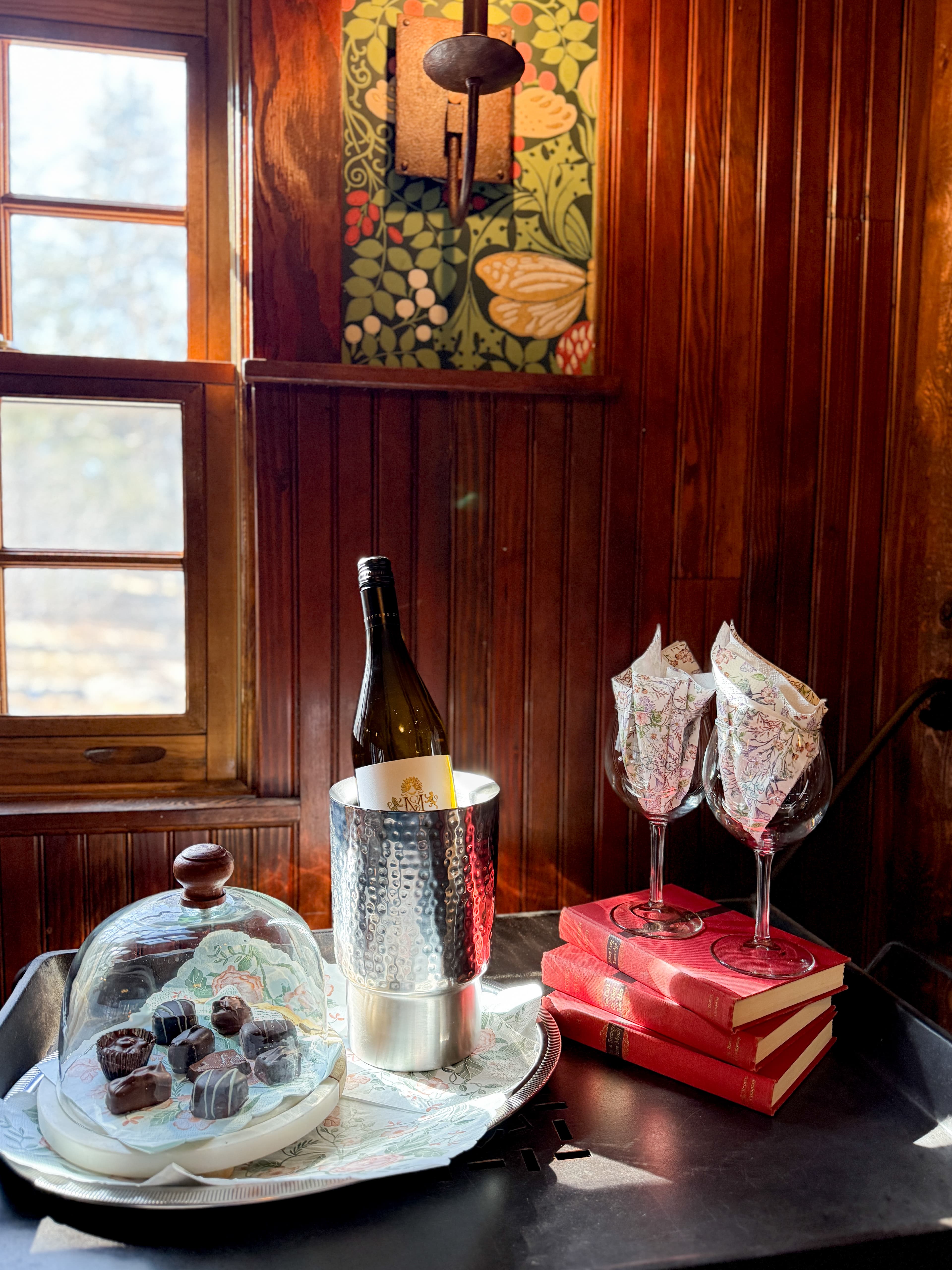 A cozy corner features a wine bottle in an ice bucket, chocolate treats under a glass dome, elegant glasses, and stacked books against a wooden wall.