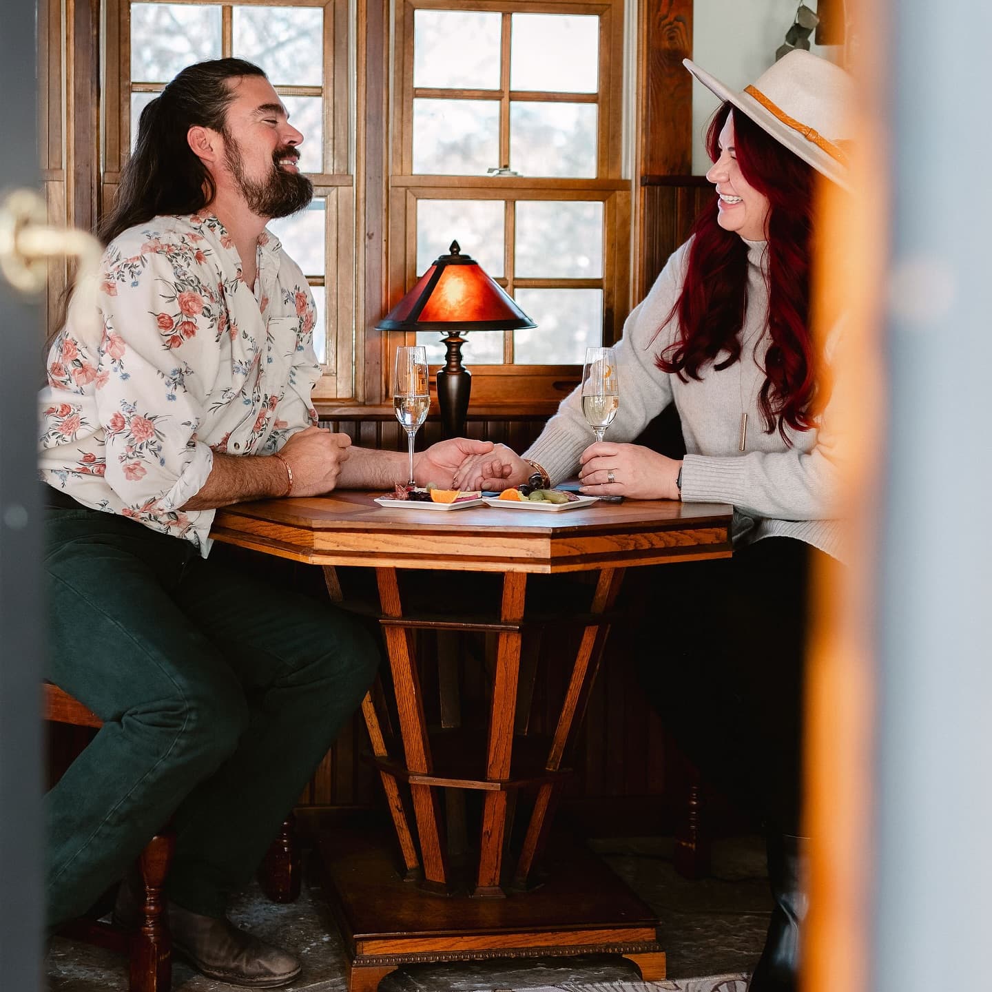 A man and a woman sit at a small table, laughing and sharing drinks in a cozy, warmly lit room.