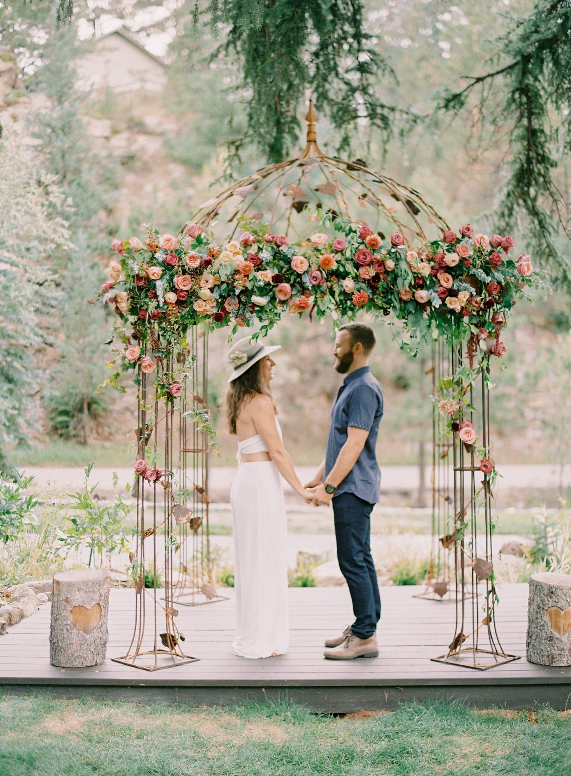 A couple stands holding hands under a floral arch in a natural outdoor setting.