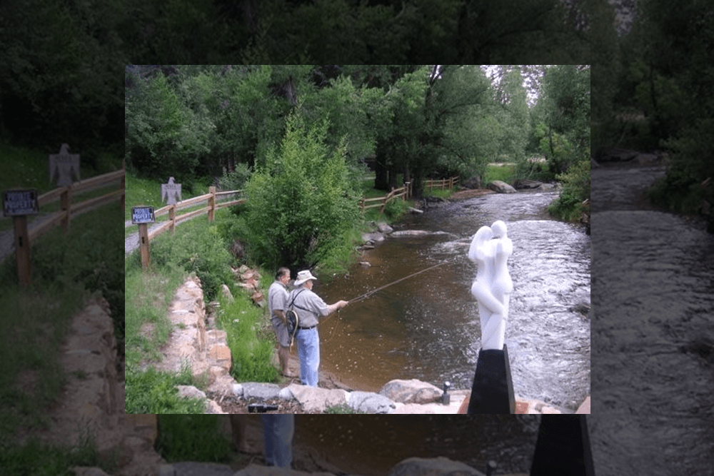Two men fish by a river, with a white statue of a couple embracing in the foreground.