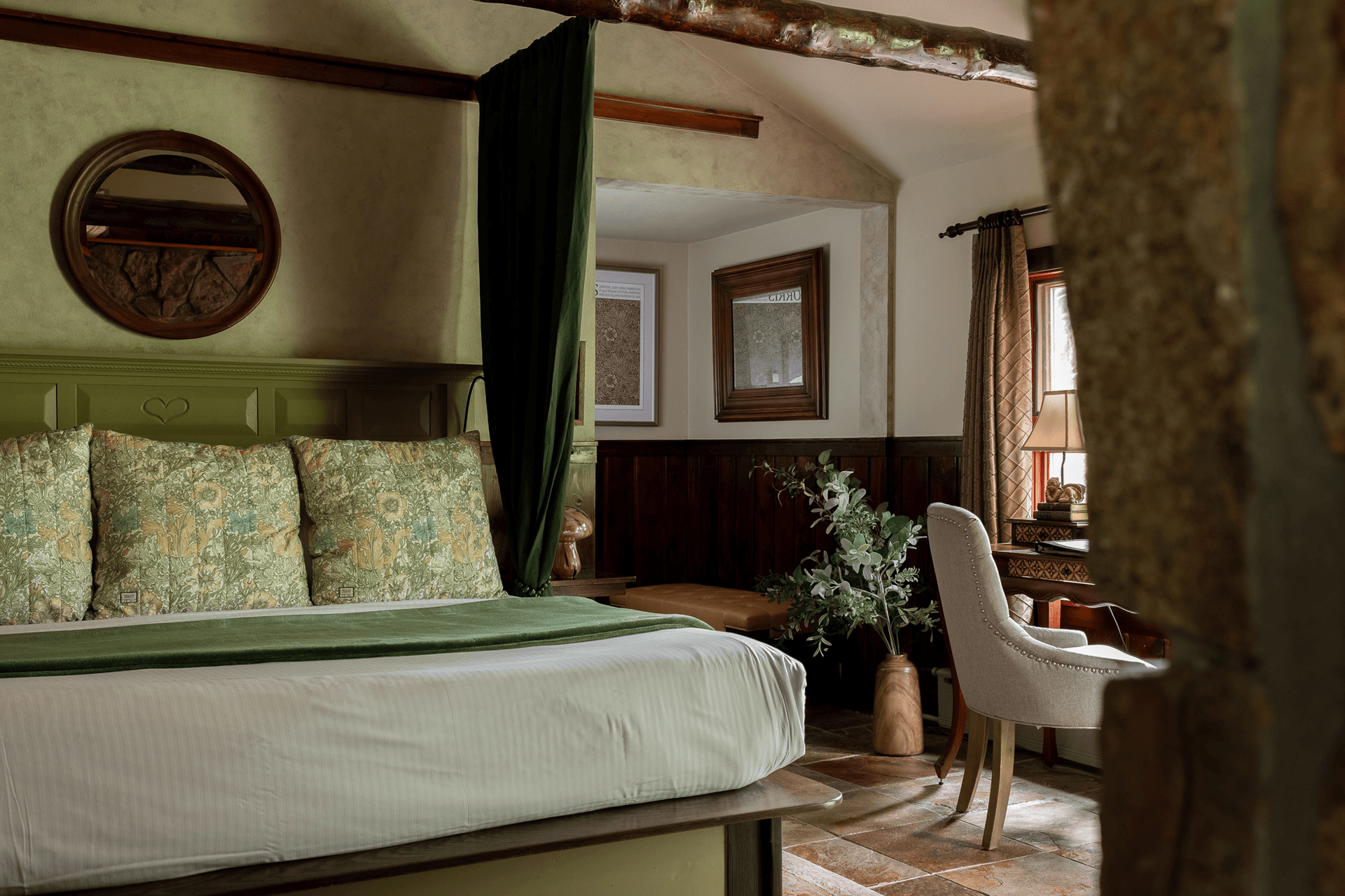 Cozy rustic bedroom with a large bed adorned with floral pillows and a green throw. Wooden beams, framed art, and soft lighting create a warm, inviting ambiance.