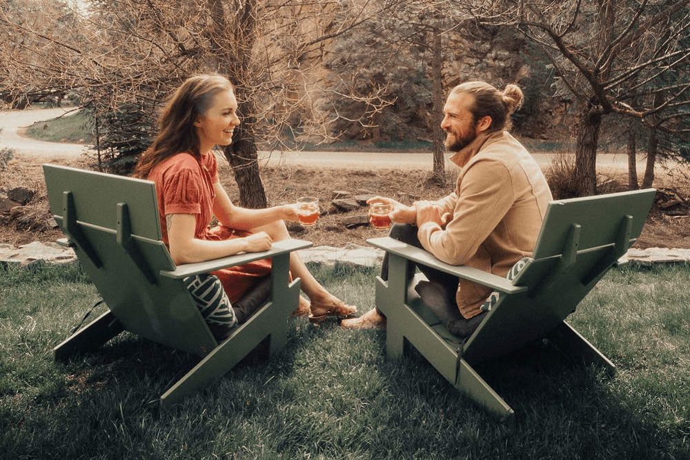 A couple sits on green Adirondack chairs, clinking glasses outdoors in a peaceful setting with trees. They're smiling, capturing a warm, relaxed vibe.
